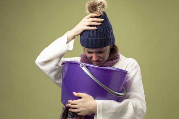 weak young ill girl wearing white robe and winter hat with scarf holding plastic bucket having nausea putting hand on head isolated on olive green background weak young ill girl wearing white robe and winter hat with scarf holding plastic bucket having nausea putting hand on head isolated on olive green background