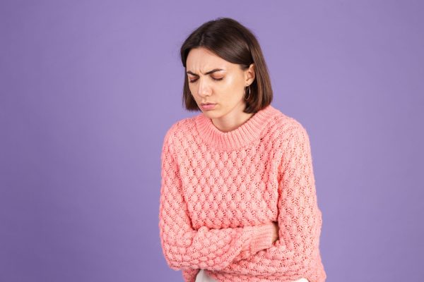 Young brunette in pink sweater isolated on purple background suffering stomach ache with painful grimace, feeling sudden period cramps, gynecology concept Young brunette in pink sweater isolated on purple background suffering stomach ache with painful grimace, feeling sudden period cramps, gynecology concept
